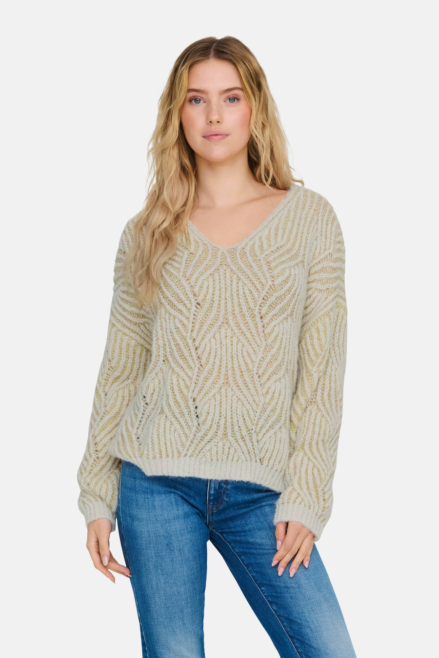 Vrouw met blonde golven in een crèmekleurige, gebreide trui met V-hals en blauwe jeans.