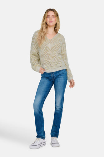 Vrouw met blonde haren draagt een beige trui, blauwe spijkerbroek en witte sneakers.