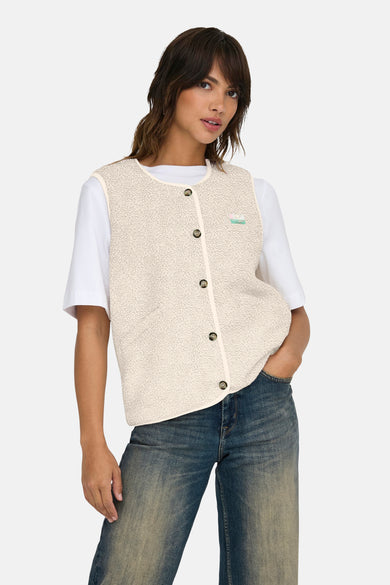 Gilet écru d'ONLY avec fermeture à boutons et un petit logo brodé, porté sur une chemise blanche et un jean bleu.