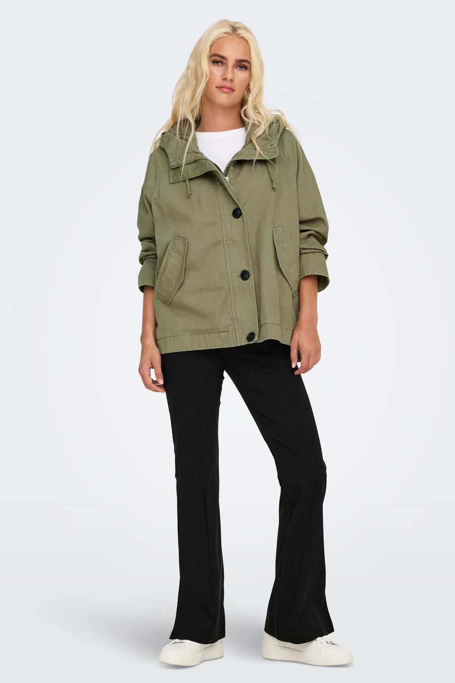 Vrouw in kaki jas en zwarte broek, staand voor witte achtergrond.