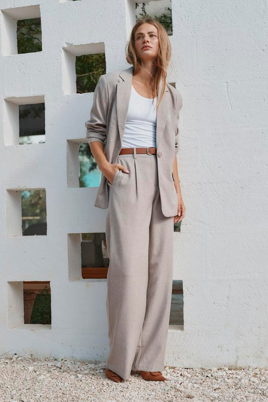 Beige kostuumbroek van ONLY® met wijde pijpen, gestikt detail en hoge taille, gedragen met lichte jas en bruine riem, lifestyle shot tegen witte muur.