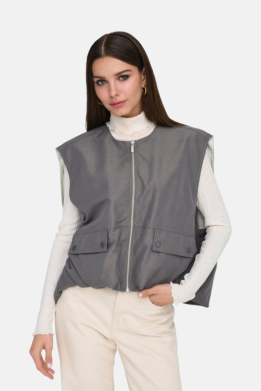 Gilet gris ONLY® avec fermeture éclair et poches, porté sur une veste blanche et un pantalon beige.
