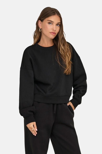 Pull-over noir avec col rond de ONLY, porté avec un short de jogging noir et des boucles d'oreilles dorées.