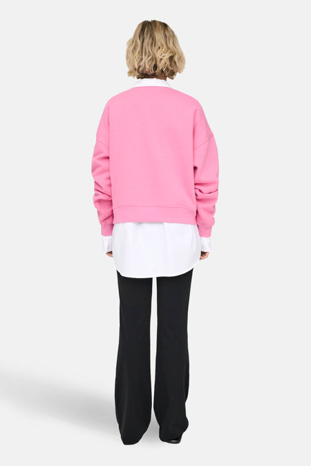 Achterkant van een vrouw met een roze trui, wit overhemd en zwarte broek.