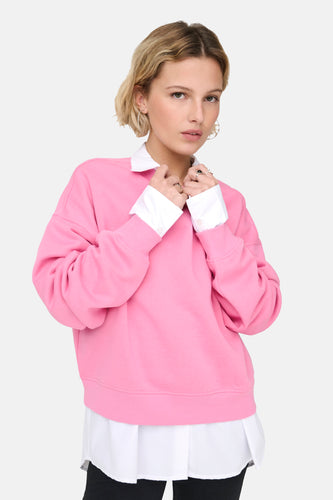 Vrouw met blond haar in roze trui met witte blouse en zwarte broek.