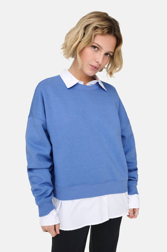 Jonge vrouw met blond haar, blauwe sweater over een witte blouse, zwarte broek op een witte achtergrond.
