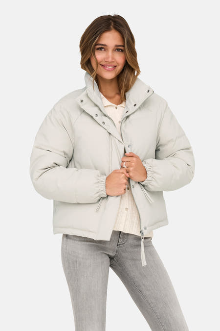 Manteau court beige de JDY, avec une fermeture éclair argentée et le modèle porte des jeans gris.