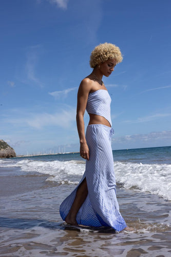 Een vrouw met blond krullend haar, gekleed in een gestreepte blauwe set op het strand.