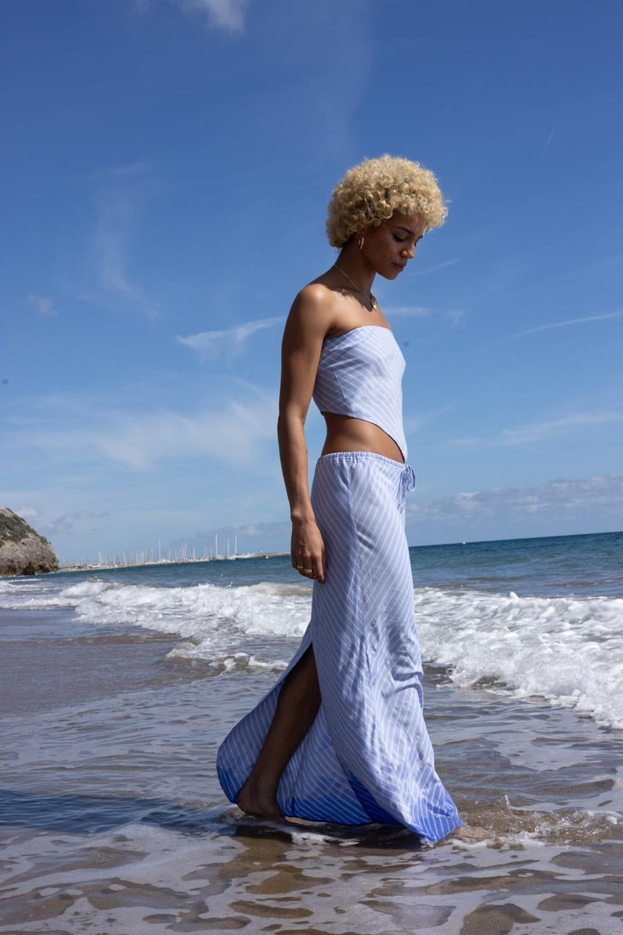 Een vrouw met blond krullend haar, gekleed in een gestreepte blauwe set op het strand.