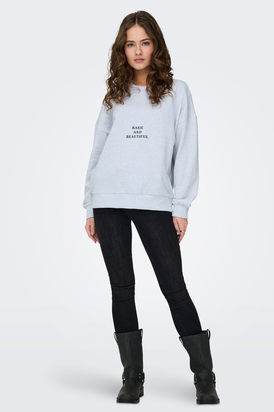 Vrouw in grijze trui, zwarte jeans en laarzen op witte achtergrond.
