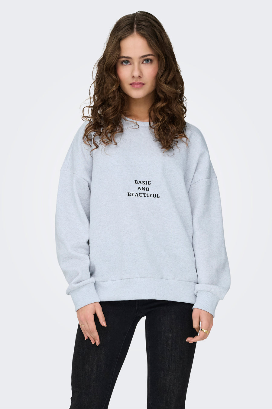 Model met krullend haar draagt grijze sweater met zwarte tekst en zwarte jeans.