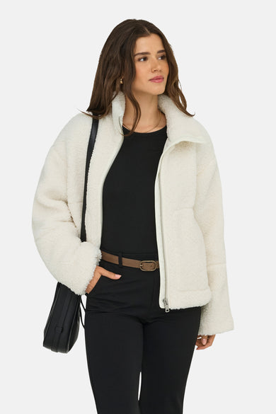 Manteau court blanc d'ONLY, avec des manches longues et une fermeture éclair avant, porté avec un haut noir et un pantalon.