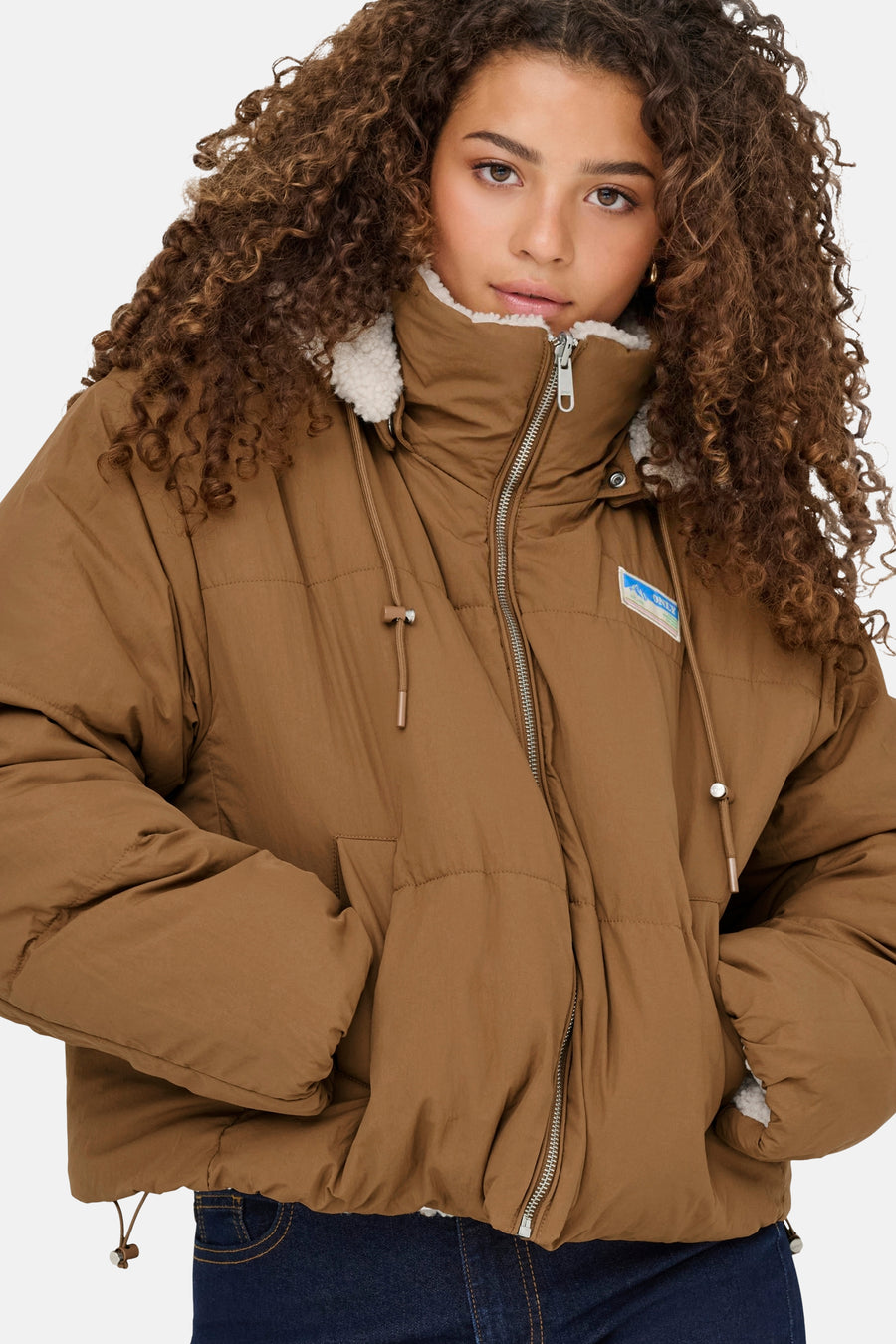 Manteau brun ONLY avec doublure en fourrure et fermetures à glissière argentées, porté avec un pantalon de cuir noir.