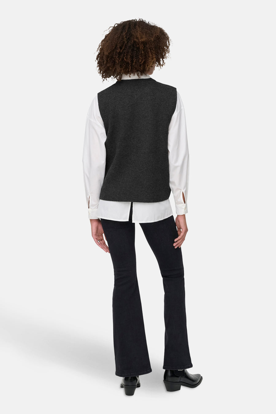 Achterkant van een persoon met krullend haar in een wit overhemd, grijze vest en donkere jeans, met zwarte laarzen.