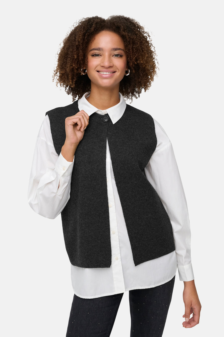 Vrouw draagt witte blouse met grijze vest en zwarte broek, lachend, op witte achtergrond.