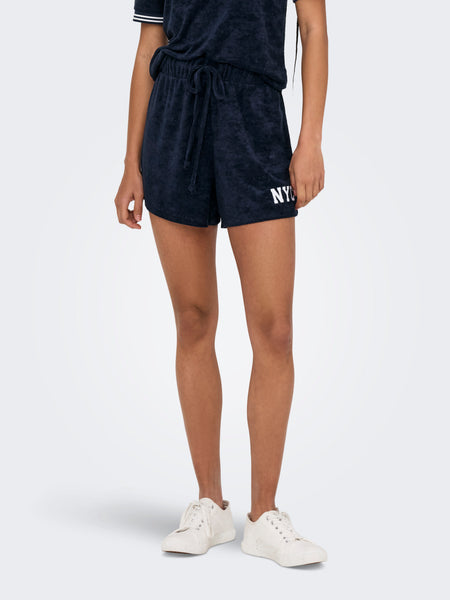 Short bleu d'ONLY, avec une ceinture et des lettres "NYC" blanches sur l'avant, porté avec des chaussures blanches.