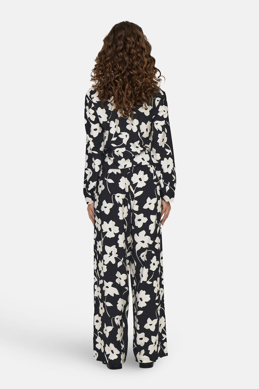 Vrouw met krullend haar, bloemenprint jumpsuit, achterkant, witte achtergrond.
