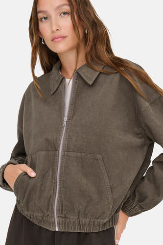 Veste courte brune d'ONLY, avec des manches longues, une fermeture éclair et des poches, portée avec un chemisier blanc.