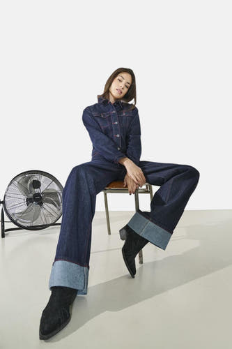 Manteau de denim bleu marine d'ONLY, porté avec des jeans larges coordonnés et des bottes noires, assis devant un ventilateur.