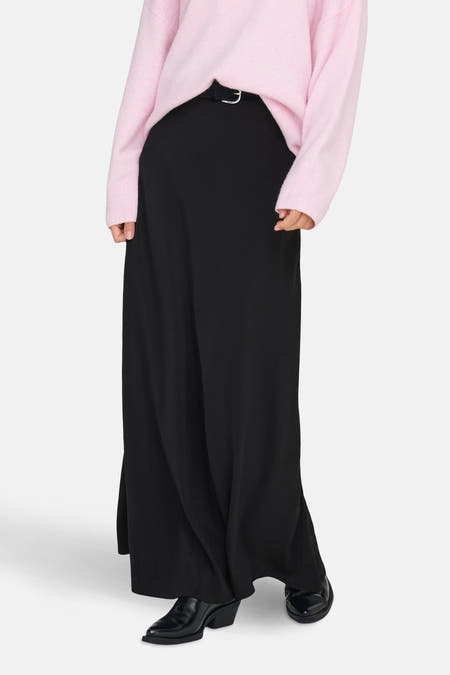 Longue jupe noire de JDY, avec une subtile ceinture et associée à un gilet rose et des bottes noires.