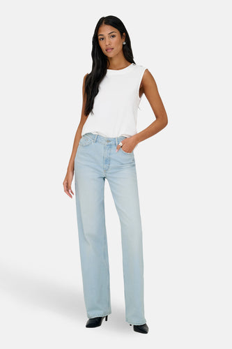 Vrouw met wit shirt en blauwe spijkerbroek staand, lichte achtergrond.