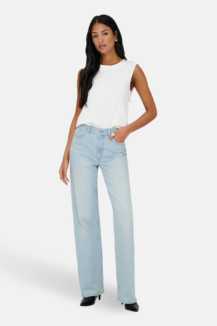 Vrouw met wit shirt en blauwe spijkerbroek staand, lichte achtergrond.