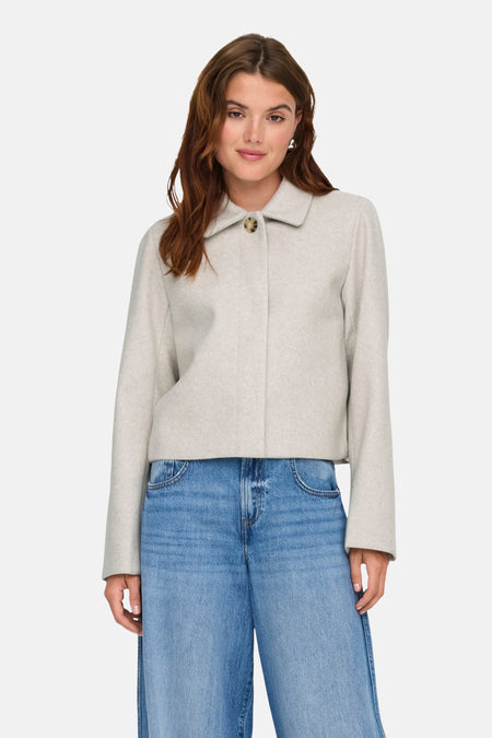 Vrouw met beige jasje en wijde jeans, staand tegen witte achtergrond.