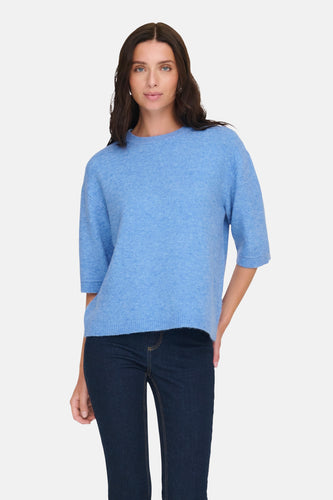 Een vrouw die een blauwe trui en donkere jeans draagt, staat tegen een witte achtergrond.