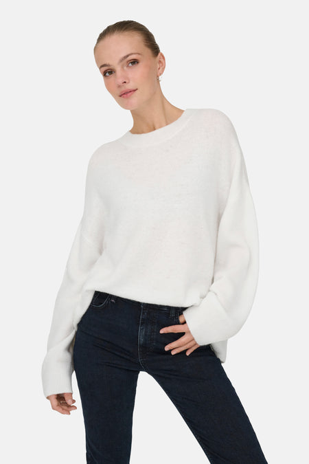 Vrouw in witte trui en donkerblauwe jeans tegen witte achtergrond.