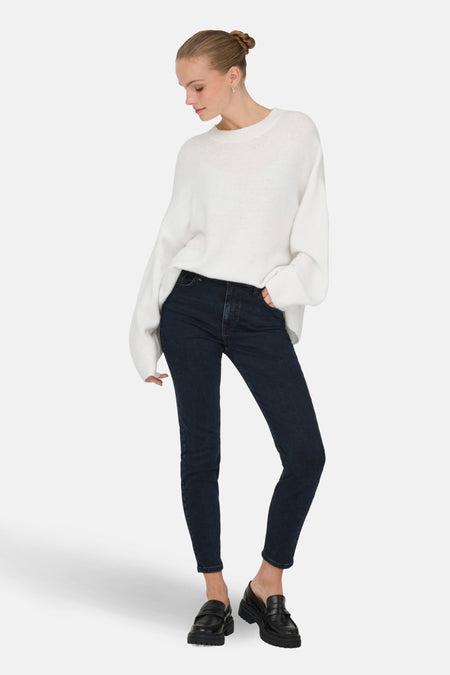 Vrouw met witte trui, donkerblauwe jeans en zwarte loafers.
