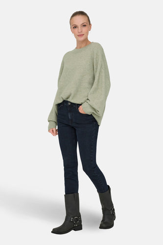 Vrouw met groene trui, donkere jeans en zwarte laarzen staand op witte achtergrond.