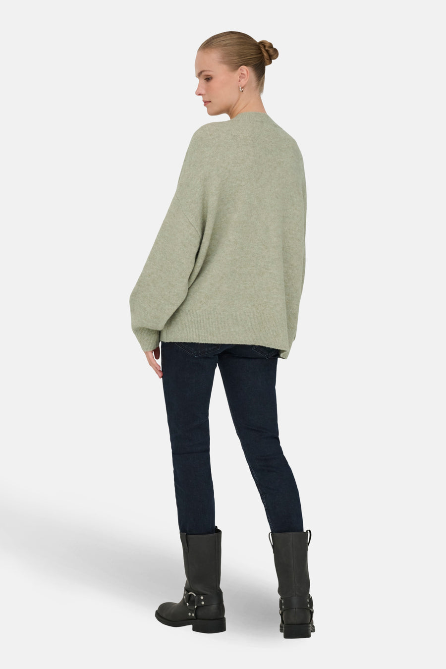 Vrouw in groene trui, donkere jeans en zwarte laarzen op witte achtergrond.