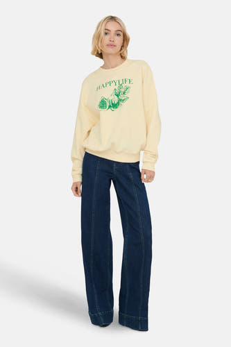 Gilet écru de ONLY avec imprimé 'Happylife' vert et image de figues. La femme porte un jean bleu marine.