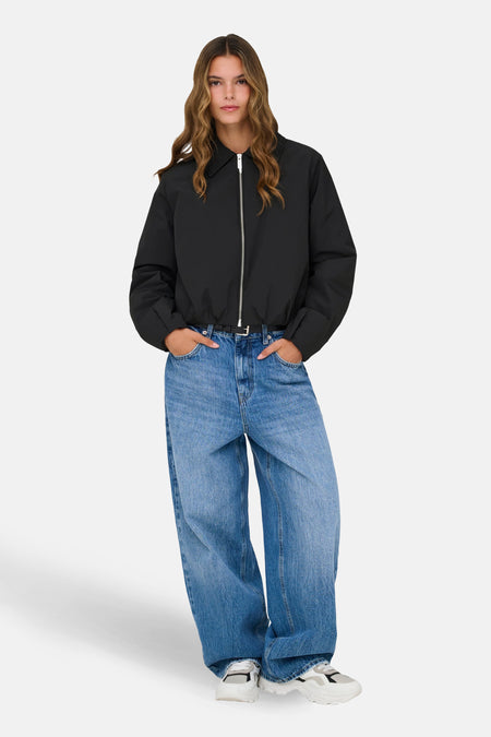 Zwarte korte jas van ONLY, met lange mouwen, een zilverkleurige ritssluiting en gedragen met wijde jeans en sneakers.
