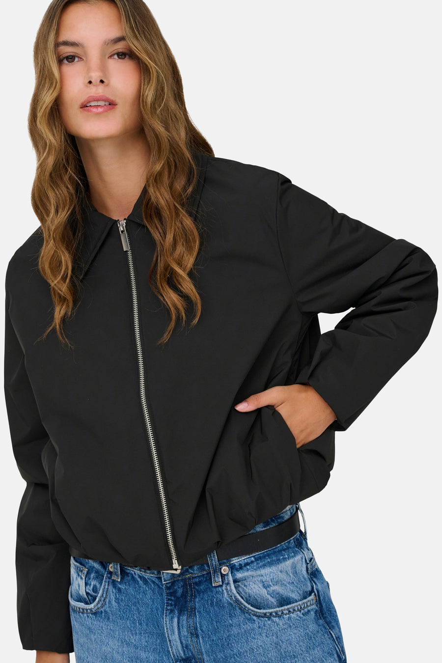 Veste courte noire de ONLY, avec une fermeture éclair argentée et une main dans la poche, portée avec un jean bleu avec ceinture.