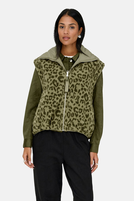 Vrouw, groene blouse, mouwloos vest met panterprint, zwarte broek, staand.
