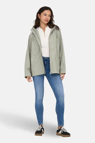Kaki regenjas van ONLY, met een capuchon, gedragen met een witte trui en blauwe jeans.
