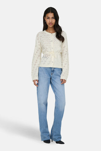 Een vrouw in een witte cardigan en blauwe jeans, staand.