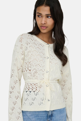 Model draagt een wit, ajour gebreid vest met lange mouwen, hartvormige knopen en een ceintuur, gecombineerd met blauwe jeans.