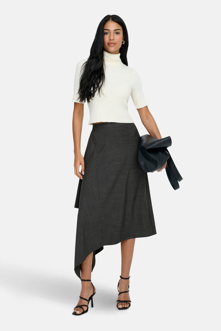 Vrouw in witte coltrui, grijze rok en zwarte sandalen met een zwarte tas in haar hand.