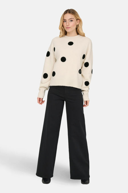 Vrouw in beige trui met zwarte stippen en zwarte broek.