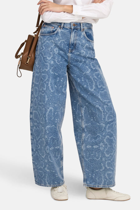 Persoon die wijde jeans met print, een wit overhemd en witte schoenen draagt, met een bruine tas over haar schouder.
