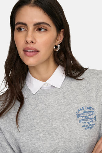 Pull gris ONLY à col rond avec imprimé bleu, porté sur une chemise blanche avec des boucles d'oreilles argentées.