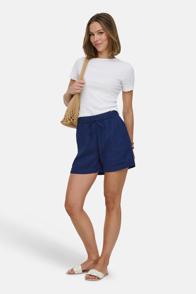 Donkerblauwe denim short van ONLY, gecombineerd met een wit shirt en een gevlochten tas, staand.

