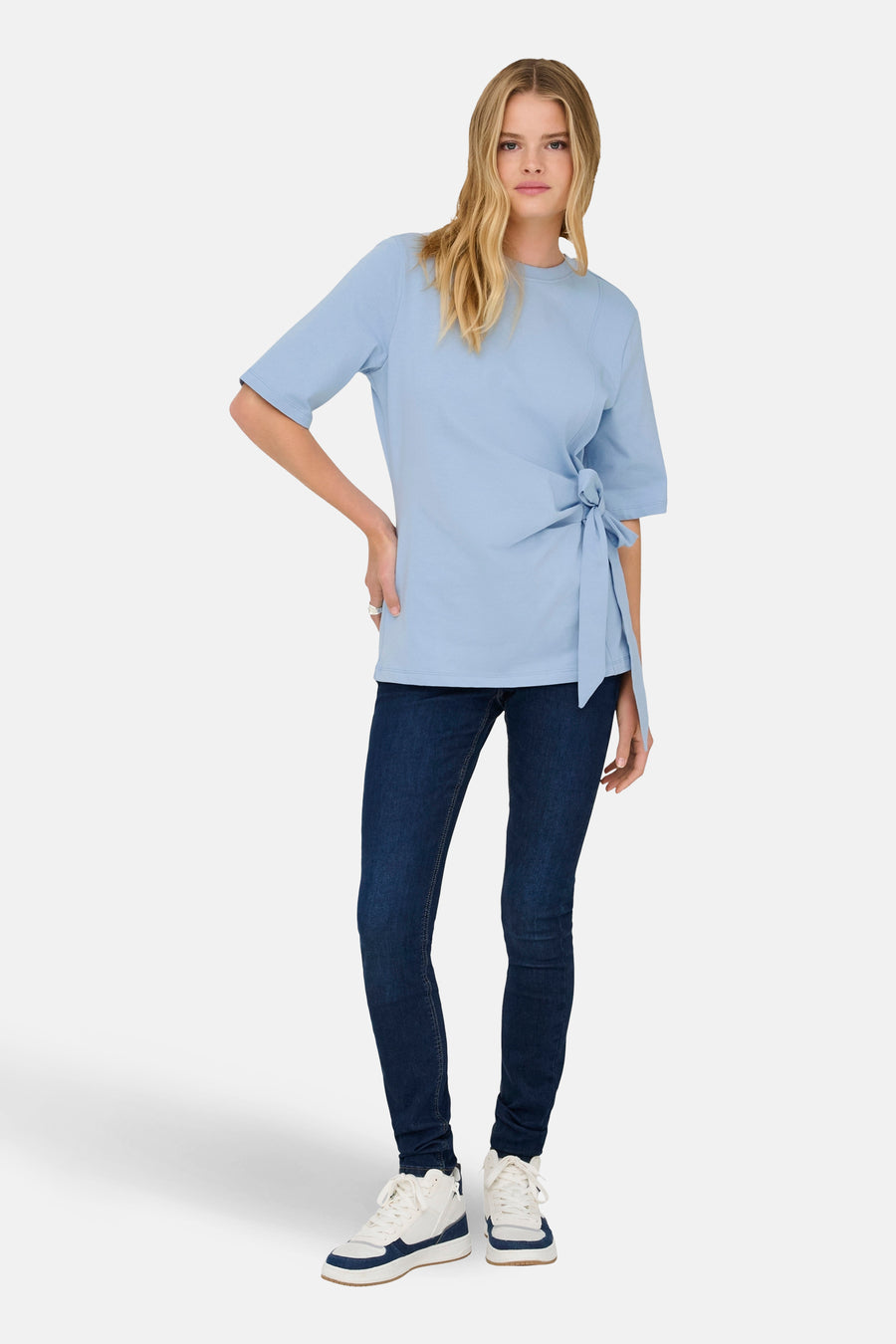 Blauw T-shirt met korte mouwen van ONLY, met een knoopdetail aan de zijkant, gecombineerd met donkere jeans en witte sneakers.
