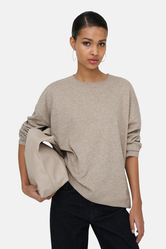 Vrouw met beige trui en zwarte broek, houdt een beige tas vast, staat tegen een witte achtergrond.