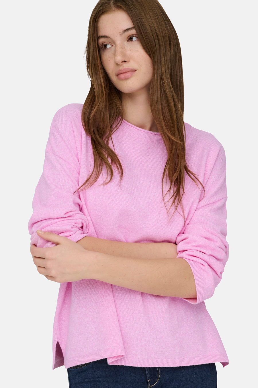 Vrouw met gekruiste armen in roze trui, staand tegen een lichte achtergrond.