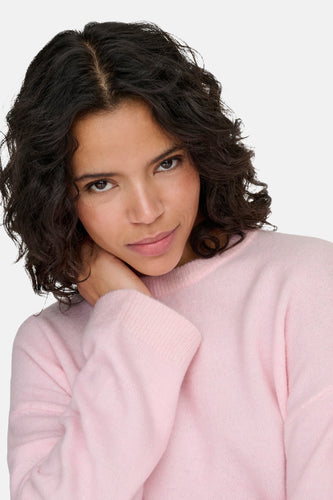 Vrouw in roze trui, hand aan nek, krullend donker haar, witte achtergrond.
