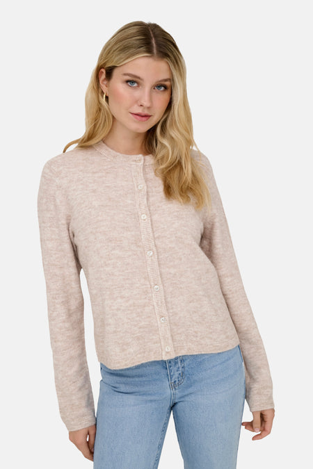 Vrouw met blonde golven in een roze cardigan met knopen en blauwe jeans.
