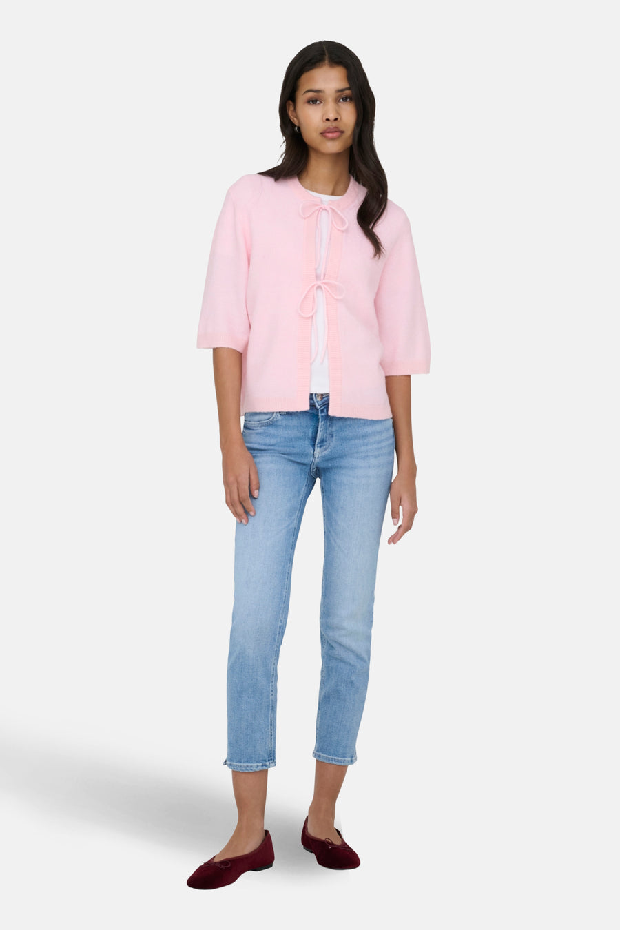 Een staande vrouw in een roze vest, een wit shirt, een blauwe jeans en rode ballerina's.